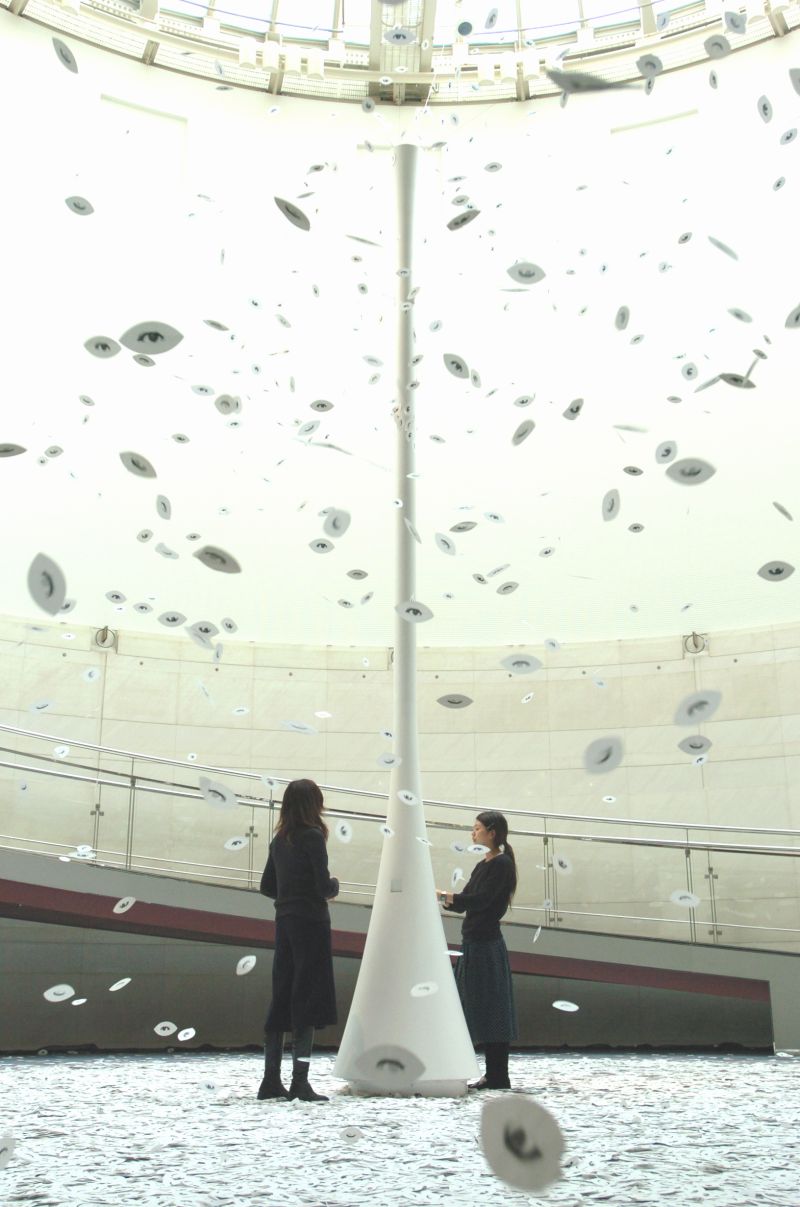 鈴木康広 《まばたきの葉》 2003年 courtesy:ワコールアートセンター photo:市川勝弘<br> <br>鈴木康広の代表作といえる参加型アート《まばたきの葉》。白い筒の先から吹き出しているのは葉のかたちをした白い紙。紙の裏表には開いた目と閉じた目をプリントされていて、紙がクルクル舞い落ちる様子はまるで葉っぱがまばたきをしているよう。筒の差し込み口から葉っぱを入れる枚数やタイミングによって、作品の表情はそのつど変わります。